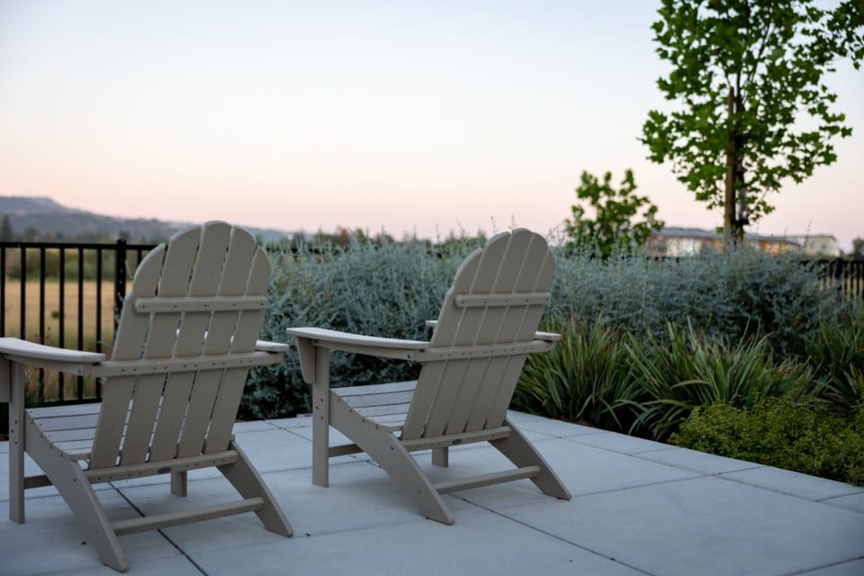 Two Adirondack chairs on a plain concrete patio beside a hedge before AI garden design