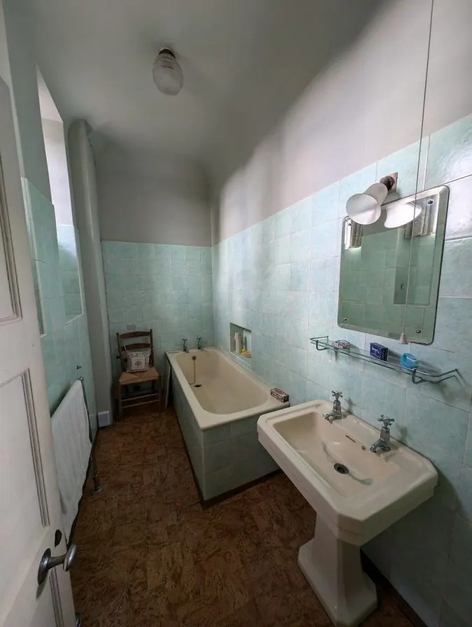 An old bathroom with pale green wall tiles, a tub, a pedestal sink and a wooden floor