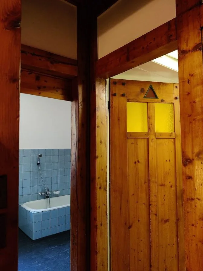 A view through wooden door frames into a bathroom with pale blue wall tiles, a tub and a dark floor