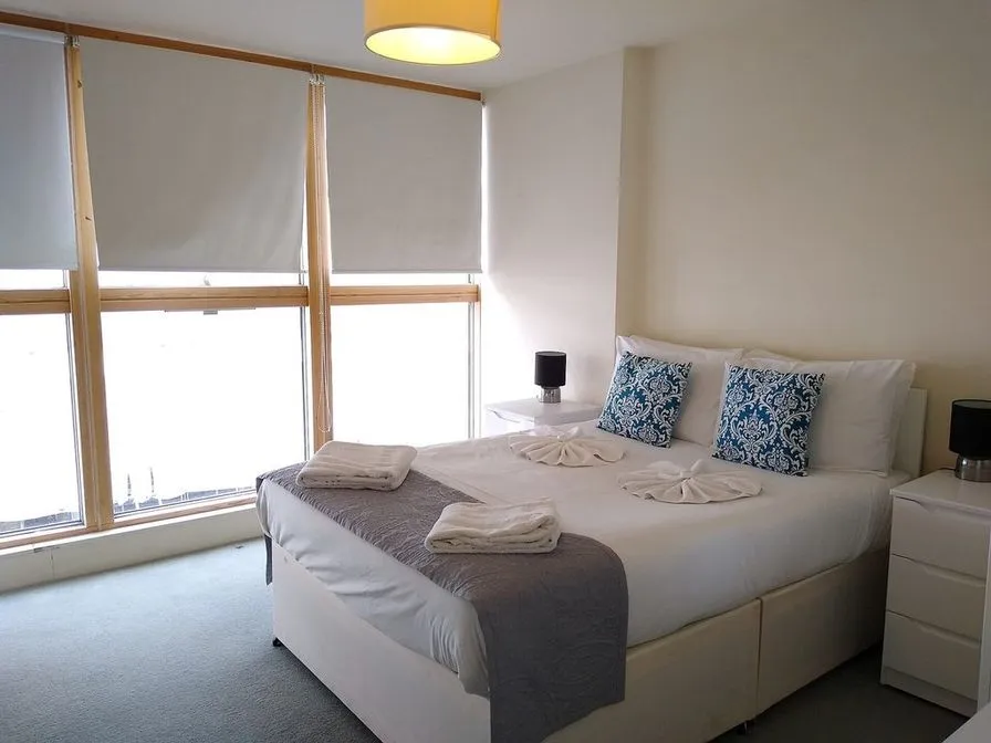 A bedroom with a double bed, blue patterned cushions, white bedside cabinets and tall windows
