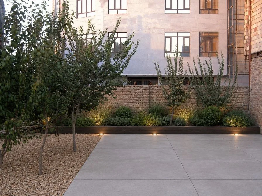 The same yard as a modern low-maintenance AI concept with large pale paving, a raised bed of grasses and low lights