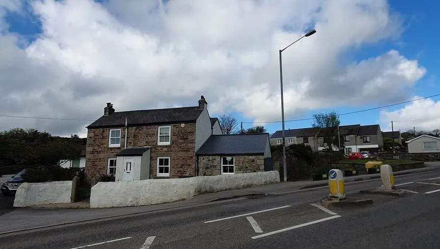 A stone and stucco cottage with a slate roof behind a low white roadside wall
