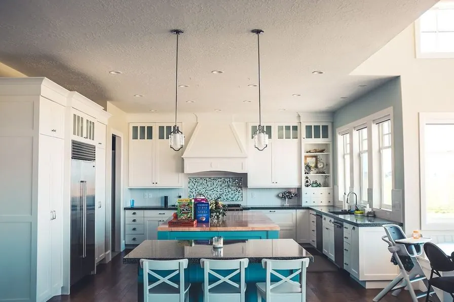 A white kitchen with an island, a teal tiled backsplash and glass pendants