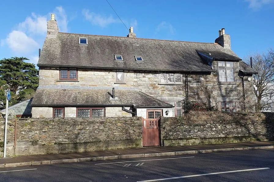 A detached stone house with a slate roof and white window frames behind a stone wall