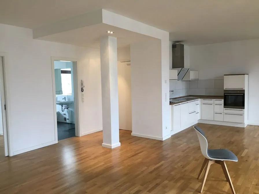 An open-plan room with a small white kitchen, a dark countertop and an oak parquet floor